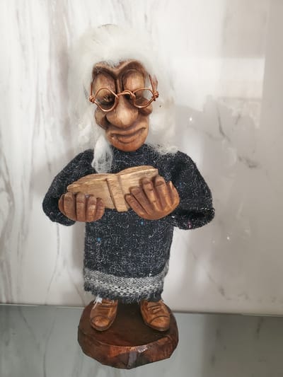 The image features a wooden figurine of an elderly person, possibly a gnome or a doll, placed on a marble-like surface. The figurine has a carved wooden face with a white beard and eyebrows. Copper wire is fashioned into glasses perched on the figurine's nose. The figure is wearing a dark, textured robe or coat with a striped cuff. Its hands are carved and appear to be holding a light-colored wooden object. The base of the figurine is also made of carved wood. The overall impression is of a rustic, handmade item. The setting appears to be indoors, with the marble surface suggesting a countertop or table. No specific time of day, weather conditions, or emotions are discernible. There is no visible text in the image. The location context provided is Kitchener, Canada, but this cannot be visually confirmed from the image itself.