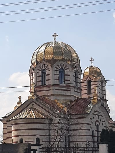 The image displays an Orthodox church edifice located in Ivanski, Bulgaria. The structure is dominated by a large, prominent golden ribbed dome topped with a golden Orthodox cross, positioned centrally. To its right, a smaller golden dome also features a similar golden cross at its apex. The church's walls are constructed with distinct alternating horizontal bands of light-colored and reddish-brown materials. Arched windows with dark blue frames are visible, notably at the base of the main dome. Sections of the roof feature reddish-brown tiles, and an additional smaller golden cross is visible on a lower roofline to the left. In the mid-ground, bare tree branches partially obstruct the lower façade of the church. The foreground includes a dark ornamental wrought iron fence. Overhead, multiple dark utility wires traverse a clear blue sky, which contains sparse white clouds. No human subjects or discernible actions are present.