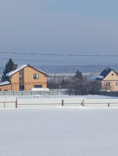 This image depicts a winter residential landscape in Яромаска, Russia, under a clear, pale blue sky. The foreground consists of a vast, unbroken expanse of white snow. In the midground, a white corrugated metal fence with dark brown posts runs horizontally, partially submerged in the snow.

Beyond the fence, two houses are visible. On the left, a two-story residence with light brown brick walls and a dark, snow-covered gabled roof stands. It features multiple dark-framed windows, including one on the upper floor and several on the lower. A dark green coniferous tree is situated to the left of this house. Behind its immediate property fence, a white, tarp-covered object, possibly a boat or vehicle, is discernible on the snow.

To the right, another house is partially visible, also featuring light-colored walls and a dark, snow-dusted gabled roof. A line of bare trees and shrubs separates the houses from the immediate foreground. In the distant background, a faint, dark line indicates a forest or land horizon, meeting the clear sky. Two dark power lines extend horizontally across the upper portion of the image. No individuals are present in the scene.