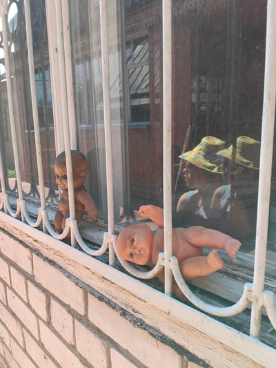 The image depicts a window with white metal bars in front of it. Through the window, two dolls are visible. The doll on the left is positioned upright, appearing to be sitting. The doll on the right is lying down, facing upwards, and appears to be a doll's head and torso. In the reflection on the window, a person wearing a camouflage hat and a light-colored shirt is visible. The building appears to be made of brick. The overall scene suggests an abandoned or neglected setting, possibly in the city of Krestovo-Gorodishche, Russia, given the provided context.