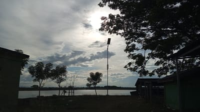 The image depicts a twilight scene with a cloudy sky over a body of water, likely a river or lake, in Keude ie Leubeue, Indonesia. Silhouetted against the bright sky are several objects: on the left, the corner of a building with a dark roof; in the center, a tall lamppost with two light fixtures; and on the right, another building with a corrugated metal roof and green walls. Various trees, also in silhouette, are scattered throughout the foreground and middle ground. The foreground is a grassy area with some scattered debris. The water's surface is calm, reflecting the muted light of the sky. The overall impression is one of a quiet, somewhat rural landscape at the end of the day.