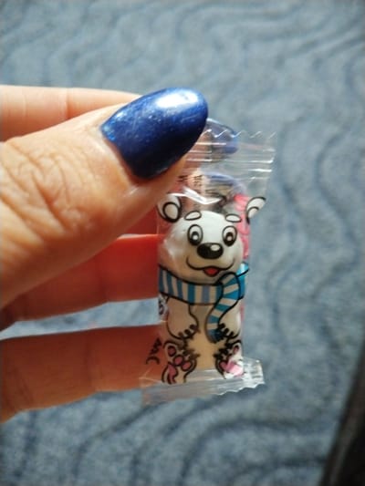A close-up, eye-level shot shows a hand holding a small, individually wrapped candy. The candy packaging features a cartoon drawing of a white polar bear wearing a blue and white striped scarf. The bear has large, round black eyes, a black nose, and a pink tongue sticking out. Its paws are depicted in pink. The hand holding the candy has a bright blue, glittery nail polish on the thumb and index finger. The background is a textured blue fabric, possibly a blanket or upholstery. The lighting is natural and appears to be coming from above, casting subtle shadows. No specific location in the city of Зелёное, Russia, is discernible from the image.