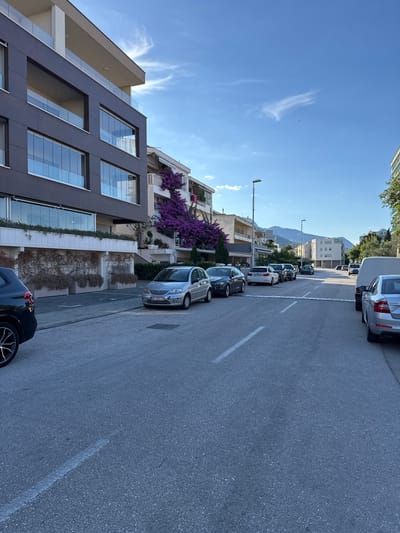 The image depicts a street scene in Makarska, Croatia. A modern, dark-colored apartment building with large glass-covered balconies is on the left. Next to it, a white building is partially covered in vibrant purple bougainvillea flowers. The street is asphalt, marked with white lines indicating parking spaces. Several cars are parked along the street, including a silver Citroen C3 in the foreground. In the background, more residential buildings and a van are visible, with mountains in the distance under a clear blue sky with wispy clouds. The scene appears to be a residential area with shops or cafes on the ground floor of some buildings.