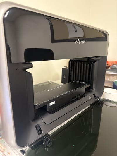 The image shows a close-up, high-angle view of an open 3D printer. The printer is black and silver, with a glossy black lid that is propped open. Inside the printer, a rectangular component with a textured surface is visible, along with a ribbed black mechanism. The brand name "eufyMake" is printed vertically in white text on the inside of the lid. The setting appears to be indoors, with a plain, light-colored wall in the background. Some office supplies, including clips and what appears to be a ruler or crafting mat, are partially visible at the bottom right edge of the frame. The lighting suggests it is daytime. No people are visible in the image.