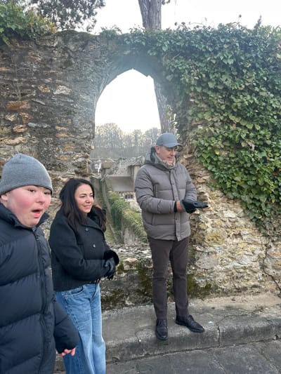 Three people pose against a stone structure partially covered with ivy. The woman in the middle wears a gray coat and jeans. The man above her wears a gray puffer jacket, brown pants, gloves, and a gray cap. The person at the bottom wears a black puffer jacket and a gray knit hat. In the background, there is a blurry view of buildings and trees. The image is rotated counterclockwise.