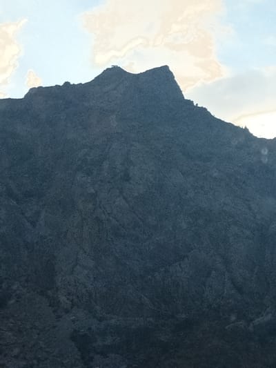 The image shows a dark, rocky mountain peak against a sky with white and light blue clouds. The mountain occupies most of the frame, with its texture appearing rough and uneven. The top of the mountain is jagged, with a sharp peak dominating the skyline. A few small, dark green trees are visible on the upper slopes of the mountain, silhouetted against the sky. The overall lighting suggests it might be late afternoon or early morning, with the sun casting shadows on the mountain. The image is a landscape photograph taken in Kurpsay, Kyrgyzstan, highlighting the natural topography of the region.
