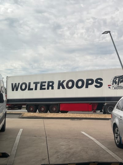 The image shows the side of a white semi-trailer truck with "WOLTER KOOPS" written in large black letters. To the right of the text is a logo with a globe and the letters "KTZ" above the word "KOOP". The truck has a red lower section and multiple wheels visible. The truck is parked on asphalt, with a light-colored vehicle to its right and the rear of another vehicle to its left, partially obscured. A lamppost is visible in the background. The scene appears to be in an outdoor lot, possibly a rest stop or industrial area, under a cloudy sky. The image was taken in Rheinbach, Germany.
