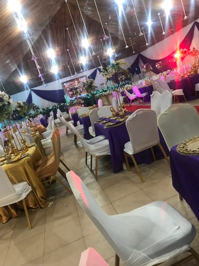 The image depicts a decorated interior space, likely a hall or ballroom, set up for an event, possibly a wedding or formal gathering. The location appears to be in Ife, Nigeria, based on a sign visible in the background.

The scene is composed of tables adorned with gold or white tablecloths and purple linens, with floral centerpieces and gold-colored decorative objects. Chairs are covered with white fabric. The ceiling is high, with decorative lights and fabric draping. No people are distinctly visible in the foreground, but figures are implied by the seating arrangements. The overall environment indicates a formal and festive occasion.