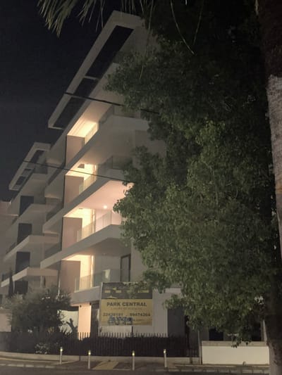 The image captures a modern, multi-story building at night, likely in Engomi, Lefkoşa, Cyprus. The building features contemporary architecture with prominent balconies or setbacks, creating a distinctive facade. Its white or light-colored exterior is illuminated by warm artificial lighting emanating from within the recessed sections and balconies, contrasting with the dark night sky.

Lush green trees and foliage are visible in the foreground and surrounding the structure, partially obscuring the building's upper levels. The low light conditions indicate it is nighttime.

On the right side of the image, a yellow and black sign is prominently displayed. The text on the sign reads: "PARK CENTRAL LUXURY RESIDENCES." Below this, two phone numbers are visible: "22438181" and "99474364." A partial web address, possibly ending in ".com," is also discernible at the bottom of the sign. No people or specific activities are visible in the scene, which appears to be a static urban landscape.