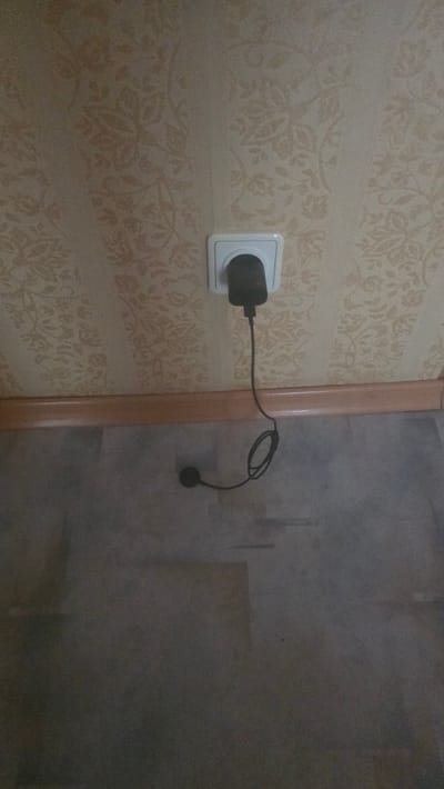 A black electrical adapter is plugged into a white wall socket on a wall with patterned wallpaper. A black cord extends from the adapter, looping onto the floor, where another black circular object is visible. The floor appears to be laminate or linoleum. A wooden baseboard runs along the bottom of the wall. The scene is likely indoors, possibly in a residential setting. There is no indication of location within Ukhta, Russia.