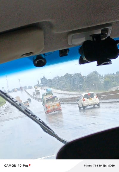 The image depicts a rainy daytime scene from inside a vehicle, looking out onto a multi-lane highway in Sigona ward, Kenya. The top section of the image shows the beige interior headliner of the vehicle, beneath which are attached a circular dark object, a black rectangular mounting bracket, and a larger black phone holder. An interior light fixture is also visible. The top edge of the windshield is tinted blue, and raindrops are present on the glass. A black windshield wiper blade extends diagonally from the lower left towards the center.
The highway, appearing wet from rain, shows traffic moving away from the viewer. In the mid-ground, a white van or minibus, marked with red and yellow livery on its rear and carrying cargo secured on its roof, occupies a central lane. To its right, in another lane, a white Honda Fit/Jazz hatchback is visible, displaying Kenyan license plate "KSD 772A". Multiple other vehicles are seen further down the highway, diminishing in size due to distance. The background consists of dense green foliage and trees lining the roadside under an overcast sky. Water spray is visible behind moving vehicles on the wet road surface.