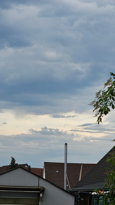 This is an outdoor, eye-level, vertical photograph taken in Vöhringen, Germany, during what appears to be overcast weather. The sky dominates the upper two-thirds of the image and is filled with a complex layering of grey and white clouds, with hints of lighter hues suggesting a distant light source. In the foreground, the rooftops of several buildings are visible. A prominent, tall, silver metal chimney stands vertically between two dark brown tiled roofs. To the left, a lighter colored building with a sloped roof is in view. On the right edge of the frame, a tree branch with green leaves and small, dark berries extends into the scene from the upper right. The overall impression is one of a rural or suburban setting under a dramatic sky.