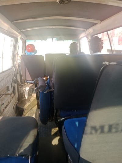 The image depicts the interior of a commuter minibus, consistent with those used for public transport in Soweto, South Africa. The perspective is from a rear passenger seat looking forward. The vehicle's interior features multiple rows of seating. In the middle row, two individuals are visible from behind: one on the left wearing a red and blue patterned cap, and another on the right with dark hair, possibly dreadlocks, and a light-colored collar. Strong sunlight illuminates the scene, causing significant lens flare. The floor is covered with a dark, patterned mat. A brown bottle and a blue object are visible on the floor between the seats in the middle row. The left side interior paneling of the vehicle appears to be removed or damaged, exposing bare metal. The seats have dark covers over blue upholstery; the back of the seat in the immediate foreground shows a faint "MER" or "MEX" imprint. The ceiling is light grey. No specific actions or interactions beyond passive seating are observed.