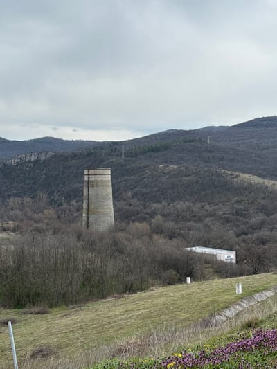An outdoor scene captured near the city of Баня, Bulgaria, depicts a landscape under an overcast sky. In the midground, a tall, weathered concrete tower, cylindrical and slightly tapered, stands amidst a dense cluster of bare deciduous trees and shrubs. To the right of the tower and lower on the slope, a white, rectangular structure, possibly a container or portable building, is partially visible, bearing the Cyrillic text "БЪД КРЕХЪК!!!" ("BE FRAGILE!!!") on its side. The background features rolling hills and mountains covered in leafless, dark-branched trees, with several lattice-structured power line pylons visible on the slopes and some rocky outcrops on the left. The foreground consists of a grassy slope, predominantly light green and brown, with patches of purple and yellow wildflowers in the bottom right corner. A concrete drainage ditch runs diagonally through the right foreground, and two white, rectangular markers are positioned on the grassy bank above the ditch. A thin, light-colored pole is partially visible on the far left edge of the foreground. No individuals, discernible actions, or interactions are present in the image.