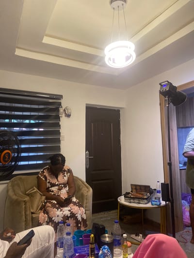 A cluttered interior in Ado-Ekiti, Nigeria. A woman in a floral dress sits in an armchair, likely using a phone. A ceiling fan stands near a window with blinds. A chandelier hangs from the ceiling. Bottles and other items clutter a table. Other people are present. A door is in the background. Lighting equipment is present.
