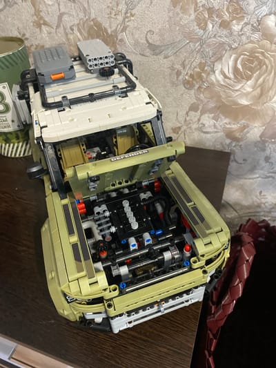 An unassembled Lego Technic Land Rover Defender is displayed on a dark wooden table, set against a wall with floral wallpaper. The model is primarily tan and white, with exposed mechanical components, including the engine, visible under the lifted hood and bonnet. Two gray Lego accessories, one resembling a storage box and the other a rocket launcher, are positioned on the roof rack. A cylindrical green object and a fragment of a green and white label with Cyrillic text are partially visible to the left of the model. To the right of the model, a segment of a dark red, woven object is present. The lighting suggests an indoor setting. The wallpaper features a textured, cream-colored background with large, embossed roses in shades of pink and cream. The overall scene is static, with no people or overt actions depicted.
