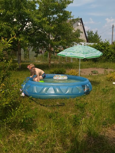 A young boy wearing orange swimming goggles and dark patterned swim trunks kneels in a blue inflatable swimming pool outdoors. The boy is positioned in the pool near its edge, with one hand resting on the side. A small, colorful inflatable ring floats in the water near him. A green patterned umbrella shades a portion of the pool. The pool is situated on a grassy lawn with trees and vegetation surrounding it. In the background, a white house with a dark roof and a satellite dish is visible, along with power lines and a greenhouse structure. The scene appears to be a residential outdoor area.