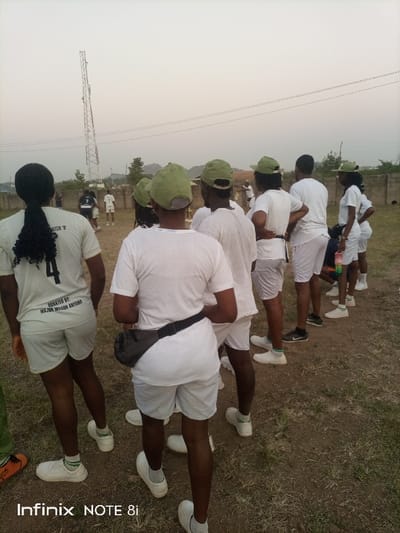 The image captures a group of young adults, likely participants in Nigeria's National Youth Service Corps (NYSC), standing outdoors in what appears to be a field or open ground. Most individuals are seen from behind, wearing a uniform consisting of white t-shirts, white shorts, and distinctive light green caps, typical of NYSC members. They also wear white socks and light-colored sneakers.

In the foreground, a person on the left has long, dark hair tied back, and their white t-shirt has the text "BATCH 'B'" and "DONATED BY: MAJ. (DR.) WILSON ANYARA" printed on the back. Another person in the mid-foreground wears a black fanny pack around their waist. The group appears to be observing an activity or event taking place further away, where a few individuals are visible in similar attire, suggesting they might be playing a game or engaged in a drill.

The setting is an unpaved area with sparse grass and dry earth. In the background, a tall, metal communication or transmission tower stands prominently against a hazy, light sky, indicative of either early morning or late afternoon. Power lines are visible stretching across the mid-ground. A low, concrete wall or fence marks the boundary of the field, with some vegetation and distant hills or buildings under the hazy sky. The overall atmosphere suggests an organized outdoor activity, possibly part of a camp or training program. A person on the far right is holding a colorful water bottle. The image includes a watermark at the bottom left that reads "Infinix NOTE 8i".