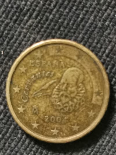 A close-up, top-down view of a single Spanish euro coin, circa 2004, is centered against a dark, textured fabric surface. The coin is golden-colored and displays the profile of Miguel de Cervantes on one side, along with the inscription "ESPAÑA" and "CERVANTES". Stars are visible around the perimeter of the coin, and the year "2004" is imprinted near the bottom. The fabric appears to be a dark gray material with a ribbed or woven pattern. The image does not depict any specific location in Bourg-lès-Valence, France.