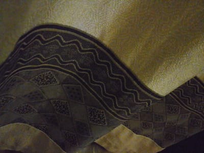 The image is a close-up, overhead shot of a textured fabric. The fabric features a wavy, dark pattern with lighter, shimmering lines against a background of varying shades of gold and green. The dark pattern is composed of multiple parallel wavy bands. Below the wavy pattern, there is a section of diamond-shaped patterns, each containing a smaller, intricate dark design. The fabric appears to be folded, creating shadows and highlights that give it a three-dimensional appearance. The lighting is warm and somewhat diffused, highlighting the texture of the material. There are no people, animals, or any discernible elements of a city or outdoor environment visible in the image. The context of Rumuomasi, Nigeria, is not visually represented in this image.
