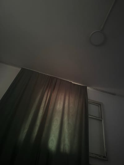 The image shows an interior view of a room, likely in Shopokov, Kyrgyzstan. The scene is dimly lit, with a large curtain dominating the left side of the frame. The curtain is a dark green color and has a subtle damask pattern visible where light hits it. The light source appears to be coming from a window partially obscured by the curtain on the right. A simple, white ceiling light fixture is visible in the upper right portion of the image, connected by a thin wire. The overall impression is one of a quiet, somewhat austere domestic space. There are no people or animals present.