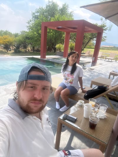 This outdoor image, captured in Pueblo Nuevo de Jasso, Mexico, depicts two individuals beside a swimming pool under a clear, sunny sky.
In the foreground, a light-skinned male with a beard and a blue baseball cap worn backward is visible from the chest up, taking a selfie. He wears a light-colored polo shirt with a contrasting collar. His lower body is partially visible, showing patterned shorts.
To the right, in the mid-ground, a female with dark, straight hair is seated on a light-brown wicker lounge chair. She wears a white t-shirt with a graphic print, blue shorts, and white ankle socks. Her posture is relaxed, with her head turned slightly towards the camera.
Between the two individuals, a low, rectangular wicker table with a glass top holds several items. These include multiple plastic cups, some containing beverages (one dark, one orange, one clear), a small green container, a small white container, a dark-colored smartphone or electronic device, and a crumpled white napkin.
Behind the female, another wicker lounge chair is visible, supporting a black bag. The swimming pool, featuring blue water and dark submerged steps, extends behind the individuals. Beyond the pool, a prominent, open-frame, rust-red architectural structure stands. The surrounding area is paved with light-colored tiles.
The background consists of lush green trees and shrubs, including some with yellow blossoms. In the far distance, low hills or mountains are visible under a partly cloudy sky, along with a building featuring a reddish roof on the right. A portion of a large, light-colored overhead canopy is visible in the upper right corner.