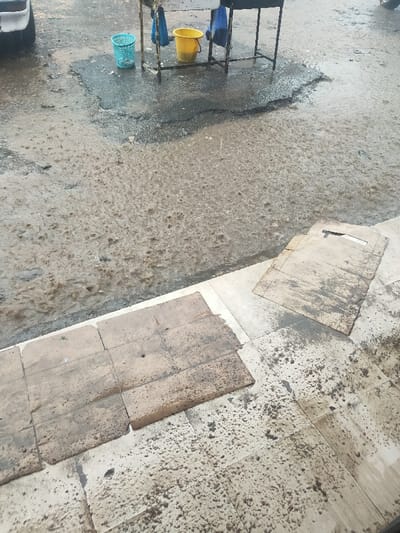 The image depicts an outdoor scene in Oloolua ward, Kenya, during a rainfall event. The ground is heavily saturated with water, showing numerous ripples and splashes from active rain.

The scene is divided into two primary ground levels: a lower, darker, uneven street or pathway covered in muddy water, and an elevated, lighter-colored paved area, likely a sidewalk or platform. The lower ground features a dark patch of what appears to be worn asphalt or dirt, completely submerged in shallow, brown rainwater.

In the upper-middle left, positioned within a large puddle, stands a dark-colored, four-legged metal framework or table. Hanging from its upper structure are two blue fabric items, possibly bags or cloths. At the base of this framework, a light blue plastic mesh basket and a yellow plastic bucket are visible, also partially submerged in water. A portion of a dark vehicle, possibly a tire and fender, is visible in the extreme upper-left corner of the frame.

The elevated paved area, composed of light-colored, possibly concrete or tiled surfaces, shows distinct water and mud splatter patterns, particularly on the tiles closer to the waterlogged lower ground. On this paved surface, two large, flattened, rectangular sheets of what appear to be wet, soiled cardboard are laid out. An additional smaller piece of cardboard is partially visible in the bottom-left corner. No individuals are present in the image.