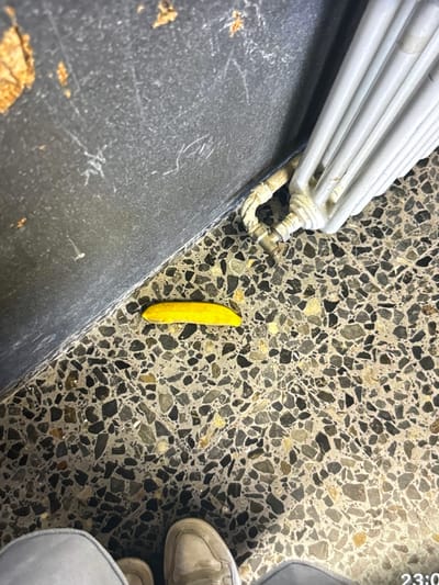 The image shows a close-up, high-angle shot of a yellow object lying on a terrazzo floor. The object appears to be a small piece of bright yellow plastic, possibly a toy or a fragment. To the left of the object is a dark grey wall, possibly concrete or painted cinder block, with some scuff marks and what appear to be flakes of rust or paint. To the right of the object is a white, ribbed structure that resembles a heating radiator or an industrial pipe. The terrazzo floor is a mosaic of grey, black, and tan stones. In the bottom portion of the image, parts of two people's shoes are visible, suggesting they are standing and looking down at the scene. The overall lighting is dim, and the focus is primarily on the yellow object and its immediate surroundings. The background is blurred.