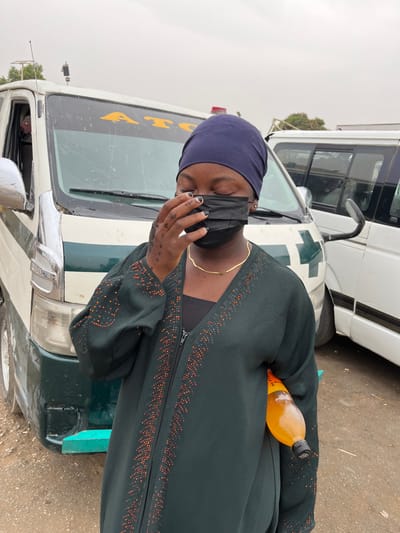 An outdoor scene in Kasuwan Kaji, Nigeria, depicts a dark-skinned individual, identified as female, standing directly in front of a white utility van under an overcast sky. The individual wears a dark blue head covering, a dark green long-sleeved abaya with patterned orange-brown circular embellishments along the zipper and sleeves, and a black top beneath. A thin gold-colored necklace is visible. Her eyes are closed, and her left hand, featuring dark nail polish and visible mehndi patterns, holds a black disposable surgical mask over her nose and mouth. An orange-colored beverage in a clear plastic bottle is tucked under her right arm.
The primary vehicle behind the individual is a white van with a prominent green horizontal stripe across its front and a large green cross symbol on the passenger side. Yellow "ATC" lettering is visible on the windshield, and a teal-colored front bumper is present. A roof-mounted antenna is also visible on this van. A second, partially obscured white van is parked to the right and further back. The ground beneath the individual and vehicles appears to be an unpaved, damp, light-colored surface.