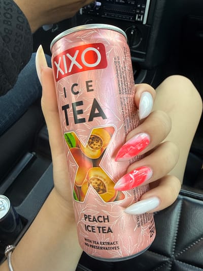 An individual's hand with long, pointed, manicured nails, holding a pink can of Xixo Ice Tea, is visible in the foreground. The can features an image of peaches and text indicating "Peach Ice Tea" and "With Tea Extract. No Preservatives." The background shows a dark interior, possibly a car, with hints of the vehicle's dashboard and seating. Another beverage can is partially visible on the left. The scene appears to be in Brezovo, Bulgaria.