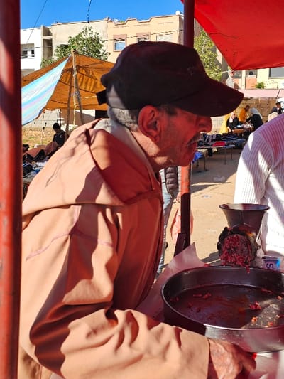 The image depicts an outdoor market or street scene under a clear blue sky. The main subject is a man, seen from a side profile, wearing a dark baseball cap and a light brown or tan long-sleeved jacket. He has stubble on his face and appears to be looking forward or slightly upwards.

To the right of the man, a manual metal meat grinder is in operation or has recently been used. Ground meat, reddish in color, is visible in the grinder's mechanism and collected in a large, shallow metal tray positioned next to it. A person wearing a white garment or apron is partially visible behind the meat grinder, suggesting they are involved in the meat preparation. A small white bottle with a blue cap, possibly containing water or another liquid, is visible near the grinder, with a faint "S" visible on its label.

The scene takes place on unpaved ground. In the background, other individuals are visible, some gathered around what appear to be market stalls. A large brown or tan fabric canopy provides shade over a section of the market. Beyond the market activity, several multi-story buildings with windows can be seen, indicating an urban or semi-urban setting. The bright sunlight casts distinct shadows, suggesting it is daytime. The foreground is partially framed by red metal bars or structures, possibly part of a stall or barrier.