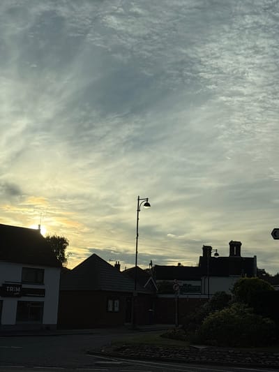 The image shows a street scene in South Staffordshire, United Kingdom, under a cloudy sky with the sun partially visible through the clouds, creating a soft, diffused light. On the left, a building with a dark roof and white facade displays a sign that reads "TRIM SHOPS" with "trimbarbershops.co.uk" below it. To the right of this building is a smaller structure with a dark, pyramid-shaped roof. Further in the background, several buildings with chimneys are visible, silhouetted against the sky. A street lamp with a globe is positioned between these buildings. In the foreground, the edge of a road with a stone curb is visible. The scene is dominated by the sky, which is filled with various cloud formations. There are no people visible in the image.
