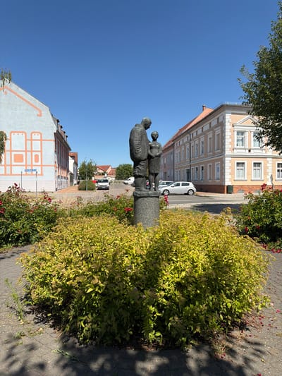 This image depicts a street scene in Schwedt/Oder, Germany. The focal point is a statue of three figures, appearing to be adults and a child, rendered in bronze. They are positioned on a stone pedestal. To the right of the statue is a lush, green bush with yellow foliage and scattered red flowers. The background features a street lined with buildings, one of which is light beige with white trim and architectural details. A white car is parked on the street. Further down the street, more buildings and trees are visible under a clear blue sky, suggesting it is daytime with fair weather. The perspective is slightly angled, with the sky occupying a significant portion of the upper part of the image. A street sign is visible on the left side, reading "Neuerdamm". The overall impression is a quiet, sunny day in a European town.