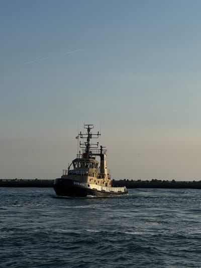 A tugboat named "DORADO" is moving through choppy, dark blue water towards the right side of the frame. The tugboat is light-colored with a darker hull, and a prominent superstructure with antennas and lights. A breakwater made of large, dark concrete blocks extends horizontally across the middle ground of the image, behind the tugboat. The sky is a pale, gradient blue, with two faint contrails visible in the upper left quadrant. The overall lighting suggests it is either early morning or late afternoon. This scene depicts the harbor entrance of Ravenna, Italy, a major port city on the Adriatic Sea.