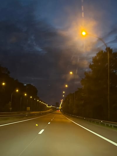 The image depicts a dark, cloudy sky over a highway at dusk or night. Streetlights illuminate the road, which has white lane markings. Trees line the sides of the highway. The scene appears to be a typical highway environment, possibly in a rural or suburban area, given the presence of trees and the absence of dense urban structures. There are no people or vehicles clearly visible, and the focus is on the road and the atmospheric conditions. The provided location of Mondercange, Luxembourg, suggests a European context for this highway scene.
