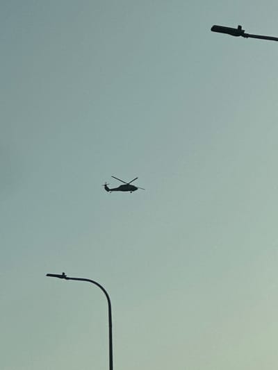 The image displays a utility helicopter in flight, silhouetted in black against a uniformly light blue, clear sky. The helicopter, which appears to be a Black Hawk variant, is positioned slightly to the left of the image's horizontal center, with its main and tail rotors distinctly visible. In the foreground, two black, curved street light poles are partially visible as silhouettes: one enters the frame from the bottom-left edge, curving upwards, and the other from the top-right edge, curving downwards. No individuals or specific ground features are discernible in the frame. The scene is located within the Galil Elyon Regional Council, Israel.