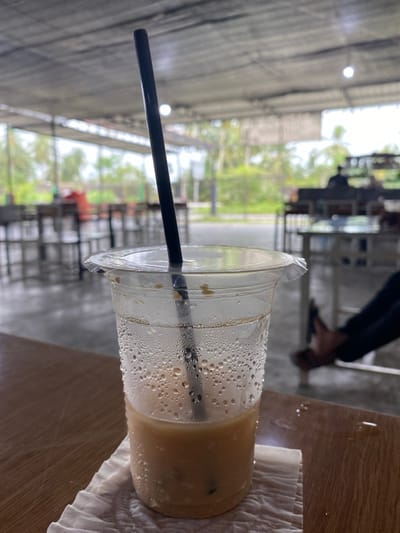 A partially consumed iced beverage in a clear plastic cup, containing a black straw and ice cubes, rests on a white paper napkin on a light brown table. Condensation is visible on the cup's exterior, and water droplets are present on the table surface. The scene is located within a semi-open commercial establishment in Aluebutuha, Indonesia, featuring a grey corrugated roof supported by metal beams and a concrete floor. Multiple light-colored tables and chairs are arranged throughout the space. In the middle-ground, the lower legs and feet of an individual, wearing dark pants and sandals, are visible to the right. A blurred figure is seated at a table in the background. Beyond the structure, a road runs parallel to lush tropical vegetation, including palm trees.