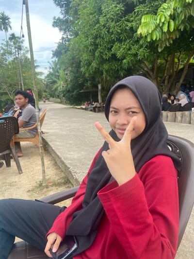 A portrait-oriented, slightly tilted photograph captures a young woman in the foreground, posing for the camera. She is wearing a black hijab and a red long-sleeved shirt. Her right hand is raised, fingers forming a peace sign. Her expression is bright and engaging. She is seated in a dark plastic chair. Behind her, a paved walkway is visible, bordered by lush green trees and foliage. In the background, other people are seated, some on steps, suggesting a gathering or outdoor event. The lighting indicates it is daytime, with bright sunlight filtering through the leaves.
Further in the background, another young man is seated in a wooden chair, also looking towards the camera. He is wearing a grey t-shirt and shorts. The scene appears to be outdoors in a tropical or sub-tropical environment, given the abundance of greenery and palm trees visible. The overall atmosphere is casual and relaxed, suggesting a social gathering or outing. No text is visible in the image.