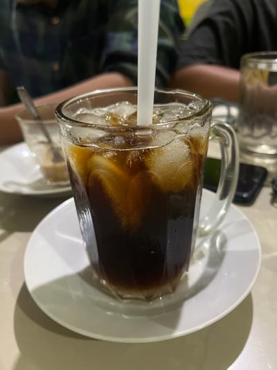 This media file shows a close-up of a glass mug filled with a dark liquid and ice, likely a beverage like iced coffee or cola, resting on a white saucer. A white straw extends from the mug. The background is blurred, but hints at a casual setting, possibly an eatery or cafe. In the background, a person's arm and shoulder are visible, suggesting they are holding another glass or item. Another glass, partially visible in the lower left, also contains a dark liquid and ice. The lighting suggests an indoor setting, possibly during the daytime or with artificial lighting. The overall impression is of a relaxed moment, enjoying a cold drink. There is no visible text in the media file.