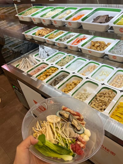 The image shows a display case filled with various food ingredients, likely for a hot pot or similar dining experience, in Bottrop, Germany. The food is organized in white rectangular containers with green trim, neatly arranged on tiered shelves. Visible ingredients include a variety of vegetables such as bean sprouts, enoki mushrooms, bok choy, and lotus root slices, as well as proteins like thinly sliced beef, mussels, shrimp, and small quail eggs. There are also processed items like crab sticks, dumplings, and various types of noodles. The display case itself is made of stainless steel and glass, with labels in German and possibly other languages indicating the food items. In the foreground, a person's hand holds a clear plastic bowl containing a selection of these ingredients, ready to be cooked. The background reveals more shelves of food, suggesting a buffet-style or self-service setup within a restaurant. The lighting is bright, illuminating the food items and the interior of the establishment.