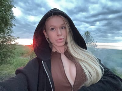 In Bolshaya Izhora, Russia, a young blonde woman with light blue eyes and a nose ring is captured in a selfie outdoors. She is wearing a dark hooded jacket zipped over a brown low-cut top. Her long blonde hair falls over her right shoulder. The background shows a cloudy sky at dusk, with a hint of sunset visible on the left horizon. Green vegetation and trees surround the area, and a body of water or distant landscape is discernible in the background. The overall impression is that of a candid outdoor portrait taken during twilight.