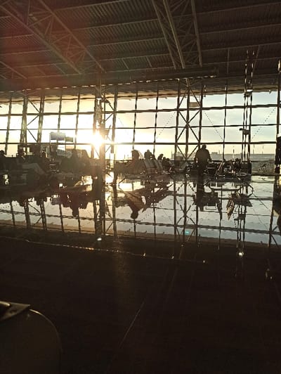 The scene depicts the interior of an airport terminal in Steenokkerzeel, Belgium, consistent with Brussels Airport (BRU). The perspective is taken from a seated position, looking towards a large, multi-paneled glass facade at either sunrise or sunset, indicated by a low sun creating a bright glare on the left side. The sky outside shows a gradient from orange-yellow near the horizon to pale blue-grey.

The terminal features a high, industrial-style ceiling with exposed metal trusses and beams. The floor is dark and highly polished, producing clear reflections of the interior and exterior light. Approximately 15-20 individuals are present, mostly seated on rows of interconnected airport-style chairs, silhouetted against the bright background. One individual is observed standing or walking towards the right side of the frame. No specific interactions or activities are discernible beyond general waiting. The overall illumination is dominated by natural light entering through the extensive glass wall.