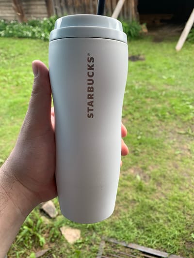 A person's hand is holding a white Starbucks tumbler outdoors, with a grassy background. The tumbler has the Starbucks logo printed vertically on its side. The background shows a wooden structure and green foliage. The scene appears to be in a rural or natural setting, likely not within a city.