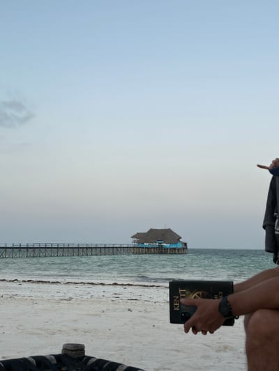 This image captures a serene coastal scene, partially showing a person relaxing on a sandy beach. On the right side of the frame, the lower torso and legs of a person are visible, holding a book with both hands. The book cover prominently displays the text "KEN TT" and "MICE," alongside a distinctive golden gear design. A watch is visible on the person's left wrist. In the upper right corner, another person's arm and hand are visible, with a finger pointing downwards.
The central and left portions of the image feature a long, wooden pier extending from the sandy shore into the calm, turquoise-blue ocean water. At the far end of the pier, a building with a traditional thatched roof and light blue walls stands on stilts over the water. The sky, which dominates the upper half of the frame, is a soft, overcast blend of grey and light blue, indicating a diffused light common to early mornings, late afternoons, or cloudy conditions. The water appears calm, and the light-colored sand of the beach shows some scattered darker elements, possibly seaweed, near the waterline. The overall atmosphere is one of tranquility and leisure, characteristic of a tropical beach setting such as Kiwengwa, Tanzania.