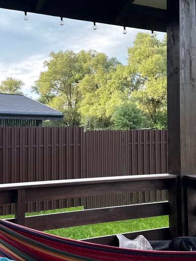 The image depicts a view from a covered porch or balcony looking out towards a fenced yard. In the foreground, a portion of a colorful striped hammock is visible. The porch features dark wooden posts and railings. Beyond the porch railing and a brown corrugated metal fence, a lush green lawn can be seen. In the background, large deciduous trees with vibrant green foliage are prominent against a cloudy sky. A dark-colored roof of a building is visible to the left of the trees. Three small, clear light bulbs are strung along the edge of the porch roof. The scene appears to be located in Gavshino, Russia, likely a residential outdoor space.