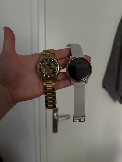 The image shows a hand holding two watches. The top watch is a smartwatch with a rose gold bezel and a light gray band. It has a blank black screen. The bottom watch is a gold analog watch with a skeleton dial, revealing the intricate mechanical workings. The background is a plain, light-colored wall, possibly an interior setting. A dark-colored folded fabric is visible in the upper left corner of the frame. The scene is well-lit, suggesting it was taken during the daytime. The focus is on the watches, implying the activity is showcasing or comparing them. No text is visible in the image. The location context of Fürstenfeldbruck, Germany is provided, but not visually indicated within the image itself.