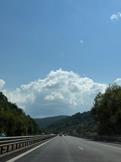 The media file depicts a roadside view on a highway in Churek, Bulgaria. The scene is captured from a moving vehicle, looking along the right lane of a multi-lane highway. The highway is flanked by a metal guardrail on the right and a steep, tree-covered hillside on the left. The sky is a bright blue with a large, dramatic cumulus cloud formation dominating the upper left portion of the image. Some smaller clouds are also visible.
Several vehicles are on the highway, appearing to be traveling in the same direction as the viewer. The overall impression of the scene suggests a bright, sunny day, possibly late morning or early afternoon, given the clear sky and the intensity of the sunlight illuminating the clouds. The lush green foliage on the hillside indicates a time of year with ample vegetation, likely spring or summer. The environment appears to be rural or mountainous, given the extensive tree cover and the absence of significant urban structures.
There is a billboard visible on the hillside with purple and white text, but it is too distant and indistinct to read. The image conveys a sense of travel and movement, capturing a scenic moment on a journey through a verdant landscape. There are no people clearly visible within the frame, only the cars on the road. The mood is serene and open, consistent with a road trip through a natural setting.