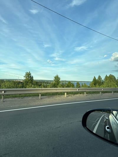 The image shows a view from a car traveling on a road, likely in or near Kondopoga, Russia, during daylight. The sky is blue with scattered white clouds and a black wire is visible stretching across the top of the frame. In the middle ground, a metal guardrail runs along the side of the road, separating it from a grassy embankment and a dense forest. Beyond the trees, a body of water is visible, possibly Lake Kondopoga or a river, and in the distance, industrial structures with smokestacks emitting smoke can be seen, indicating an industrial area within or near the city. A side-view mirror of the car is visible in the foreground on the right, reflecting the road and parts of the car.