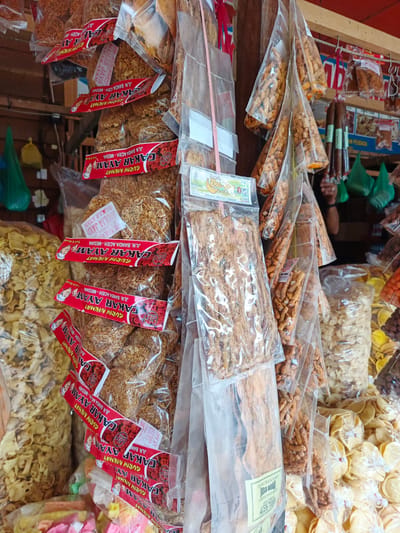 This is a medium-shot, eye-level, outdoor photograph of a market stall in Sawang, Aceh, Indonesia, selling various packaged snacks. The foreground is dominated by an assortment of clear plastic bags filled with crunchy snacks, most of which are labeled with red and white packaging. One prominent stack of snacks, labeled "CAKAR AYAM GURIH NIKMAT", features a red banner with an illustration of a smiling woman and text that includes "JL. BANCA ACEH - MEDAN" and "MULIA JAYA".
To the left, a large bag of yellow, wavy crisps is visible. To the right of the stack of "CAKAR AYAM" snacks, several triangular clear plastic bags filled with small, elongated, orange-ish snacks are hanging. Further to the right, another stack of clear plastic bags contains a variety of snacks, including some that appear to be flat, yellow chips.
In the background, more snacks are visible, including some dark, sausage-like items hanging. Green plastic bags are also scattered throughout the stall. A portion of a person's arm and shoulder is visible in the upper right quadrant of the image, indicating the presence of a vendor or customer. The stall appears to be constructed of wood and metal shelving. The overall impression is one of a bustling local market offering a wide selection of packaged food items.