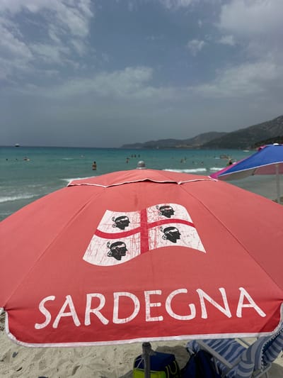 The image is a close-up shot of a red beach umbrella at the beach of Crabonaxa, in Villasimius, Italy. The umbrella has the word "SARDIGNA" written in large white letters and the flag of Sardinia emblazoned on it. In the background, the turquoise sea stretches out, with a few people swimming and standing in the water. A blue and pink umbrella can be seen to the right, and further in the distance, hills form the coastline. The sky is partly cloudy. The foreground shows the sandy beach with a beach chair and bags.